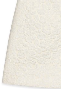 Krämfärgad tyg med ett texturerat blommönster, där upphöjda, broderade element skapar en dimensionell effekt.