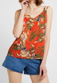 Röd blommig cami med gröna och gula tropiska mönster, med tunna axelband och V-ringning. Kombinerad med denimshorts.