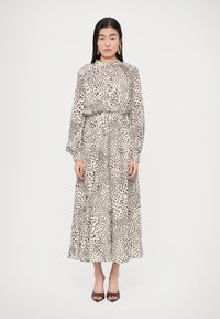 Hosszú, könnyű ruha bézs alapon fekete foltmintával, magas nyakkal, fodrozott ujjakkal és derékrészen megkötve, barna magas sarkúval párosítva.