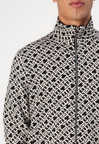 Hombre con una chaqueta de cremallera negra y blanca con un patrón de formas geométricas entrelazadas y un cuello alto, mostrado desde los hombros hasta la boca.