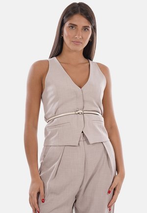 Donna con lunghi capelli scuri che indossa un gilet beige senza maniche con scollo a V e pantaloni plissettati a vita alta coordinati, in piedi con le mani rilassate ai lati.