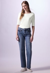 Lys grøn, tætsiddende top med trekvartærmer, parret med højtaljede, løse blå jeans. Hvide sneakers med let strukturoverflade.