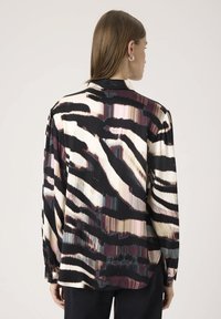 Kvinde med glat brunt hår iført en langærmet bluse med et abstrakt stribet mønster i sort, creme, pink og burgunder, vendende bort.