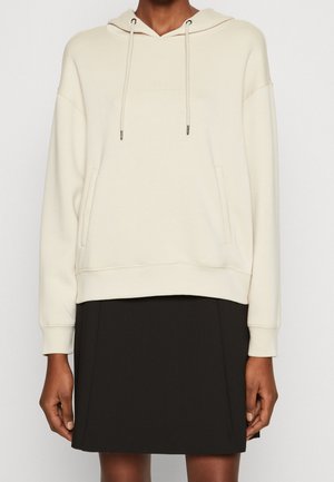 Person wearing an off-white hoodie with front pockets and drawstrings, paired with a black knee-length skirt against a plain background.
