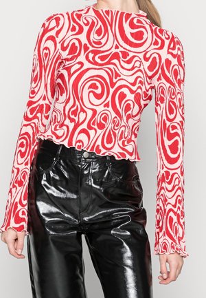 Femme portant un haut à manches longues rouge et blanc avec un motif tourbillonnant, assorti à un pantalon taille haute en similicuir noir brillant.