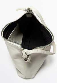 Borsa a mano in pelle beige con chiusura a zip. L'interno aperto presenta una fodera nera e una piccola tasca laterale. Dotata di un dettaglio di tracolla annodata.