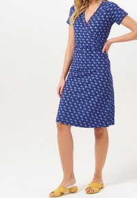 Modré šaty s obepínacím designem a krátkými rukávy, zdobené vzorem modrých vln. Na modelce jsou kombinovány s žlutými skluznými sandály.