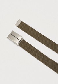 Olivno zelena tkanina pas s srebrno kovinsko sponko z gravuro "Carhartt" logotipa, položen na svetli podlagi.