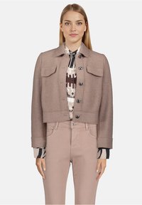 Skórzana beżowa kurtka o fakturowanej tkaninie, z zapięciem na guziki, dwoma kieszeniami na piersi i kołnierzykiem złożonym, zestawiona z wzorzystą bluzką i jasnymi spodniami.