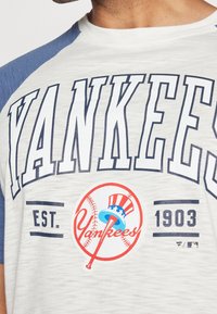 Vaaleanharmaa lyhythihainen baseball-paita, jossa tummansiniset raglan-hihat. Tässä paidassa on rohkea "YANKEES"-teksti, logo ja perustamisvuosi sinisellä.