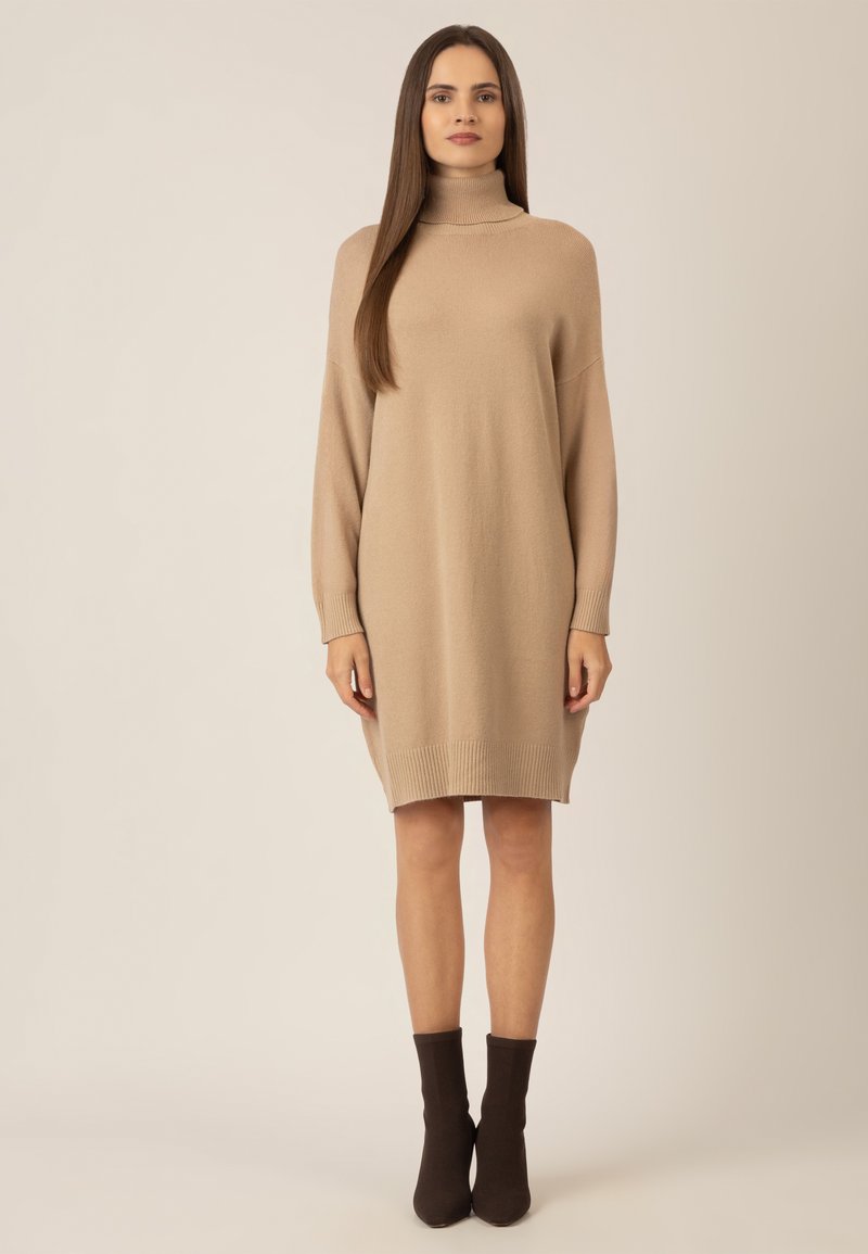 Beige gestricktes Rollkragenkleid mit gerippten Bündchen und Saum. Das Model trägt dunkelbraune Knöchelschuhe. Schlichte, lässige Silhouette.