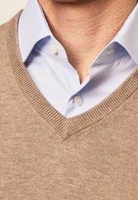 Beige knit sweater with a V-neck design layered over a light blue button-up shirt, featuring a subtle texture and ribbed edges.