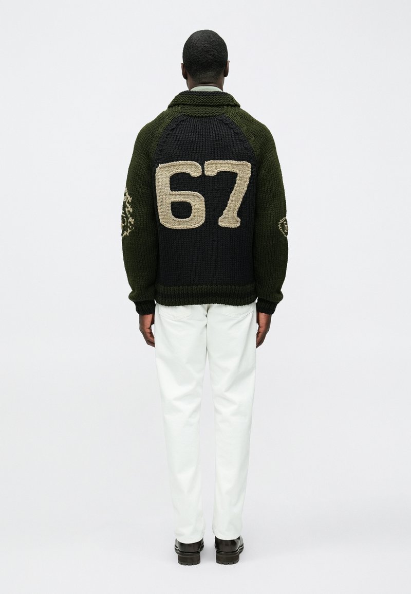 Pull en maille vert et noir avec le numéro "67" en beige dans le dos ; doté d'un col châle et d'un tissu texturé. Porté avec un pantalon blanc.