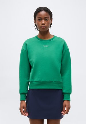 Mujer con cabello corto trenzado, vestida con una sudadera verde con el texto "30904" y una falda azul marino, sobre un fondo blanco liso.