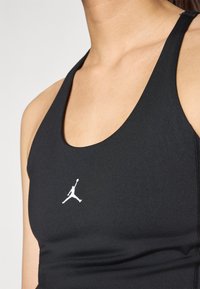Soutien-gorge de sport noir en tissu lisse, présentant une coupe ajustée et un logo Jordan blanc à l'avant. Col montant et style dos nageur.