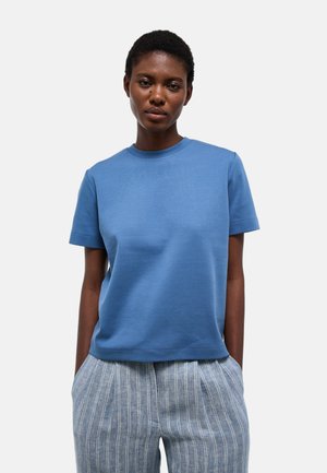 Femme aux cheveux courts portant un t-shirt bleu uni à manches courtes et un pantalon gris rayé, debout, les mains dans les poches, sur fond blanc.