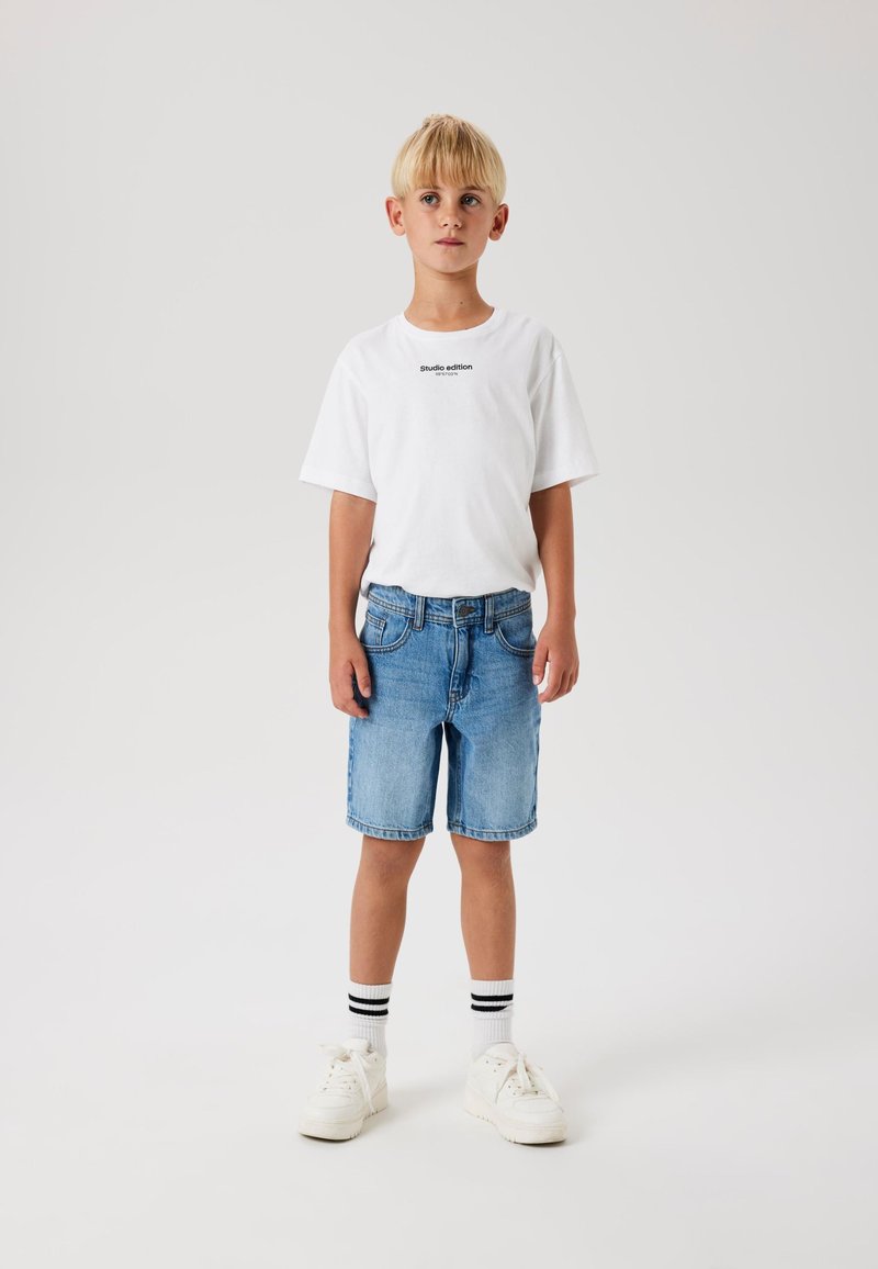 Bela bombažna majica s tekstom "studio edition", modri jeans kratke hlače in bele športne copate s črnimi črtami na črnih nogavicah.
