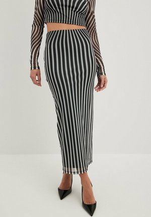 Kvinde iført sort og hvidt lodret striped lang nederdel og matchende crop top med gennemsigtige striped lange ærmer, sorte spidse hæle.