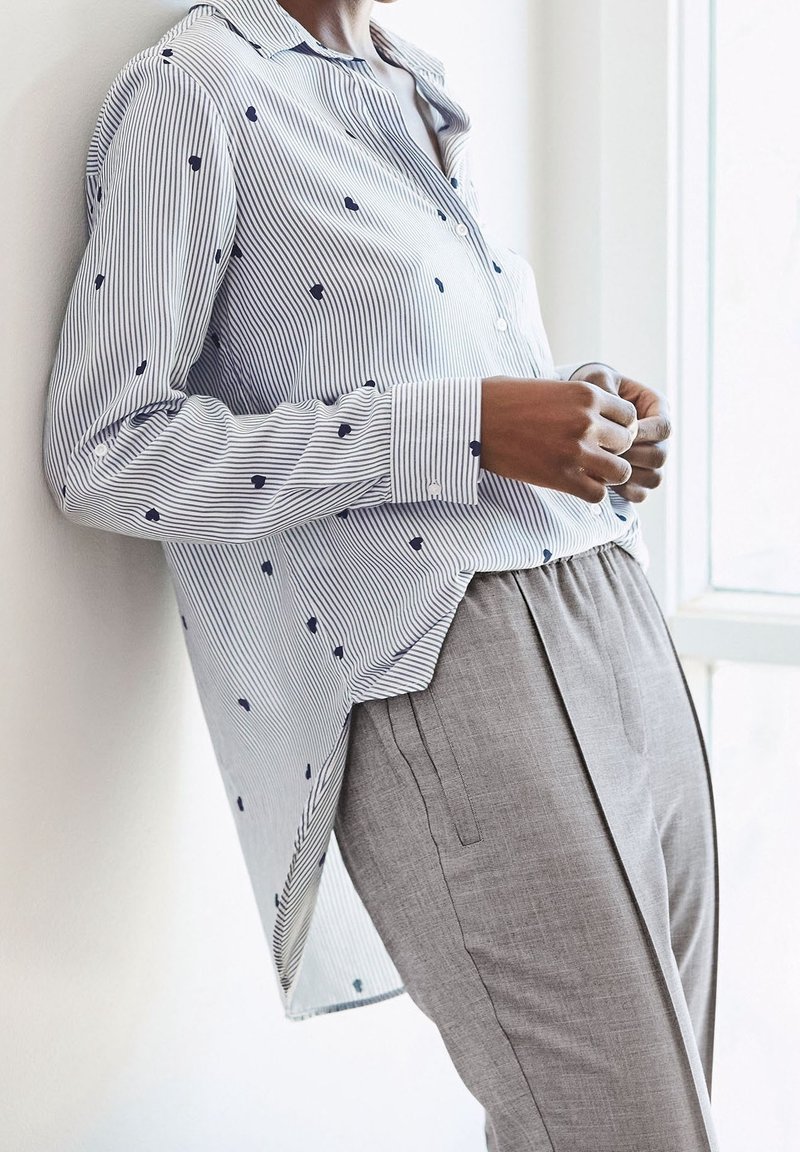 Personne portant une chemise rayée à manches longues avec de petites impressions foncées et un pantalon gris sur mesure, appuyée contre un mur blanc près d'une fenêtre.