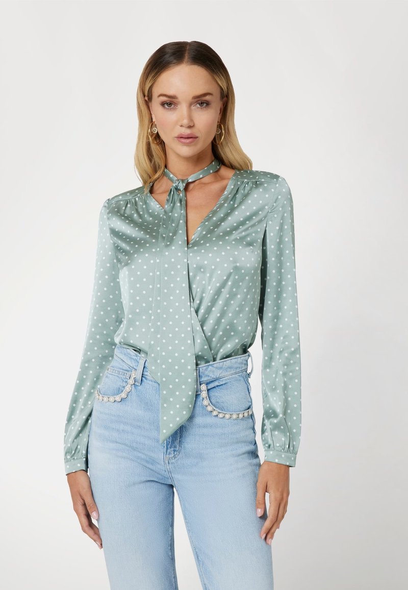 Žalia satino bluzelė su baltais taškeliais, su kaklo raiščiu ir ilgomis rankovėmis, dėvima su mėlynomis džinsinėmis kelnėmis, puoštomis dekoratyviniais akcentais.