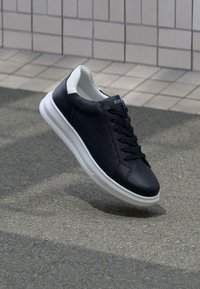 Schwarze Ledersneaker mit weißer Sohle und Schnürsenkeln, schwebend über strukturiertem Pavement und gefliestem Wandhintergrund.