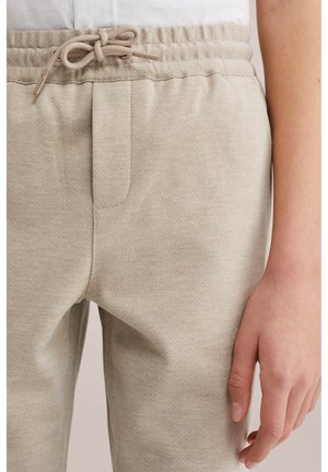 Pantaloni bej cu textură, cu talie elastică și șnur legat, purtați cu o cămașă albă băgată în pantaloni; mâna dreaptă relaxată pe lângă corp.