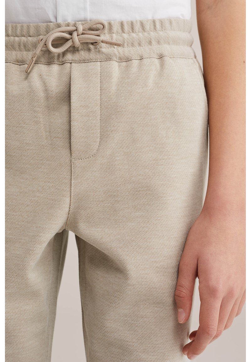 Bézs szövetnadrág elasztikus derékrésszel és megkötött húzózsinórral, fehér, belebújtatott inggel viselve; a jobb kéz lazán az oldal mellett.