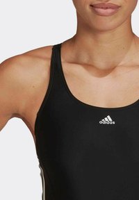 Maillot de bain de sport noir avec un design à dos nageur, comportant le logo Adidas blanc sur la poitrine. Texture lisse, matériau léger et style minimaliste.