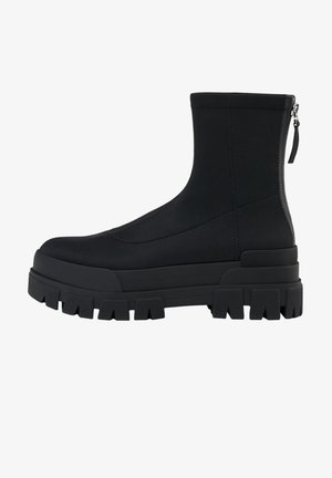 Bottine noire à semelle crantée épaisse, tige lisse et fermeture éclair arrière sur fond blanc.