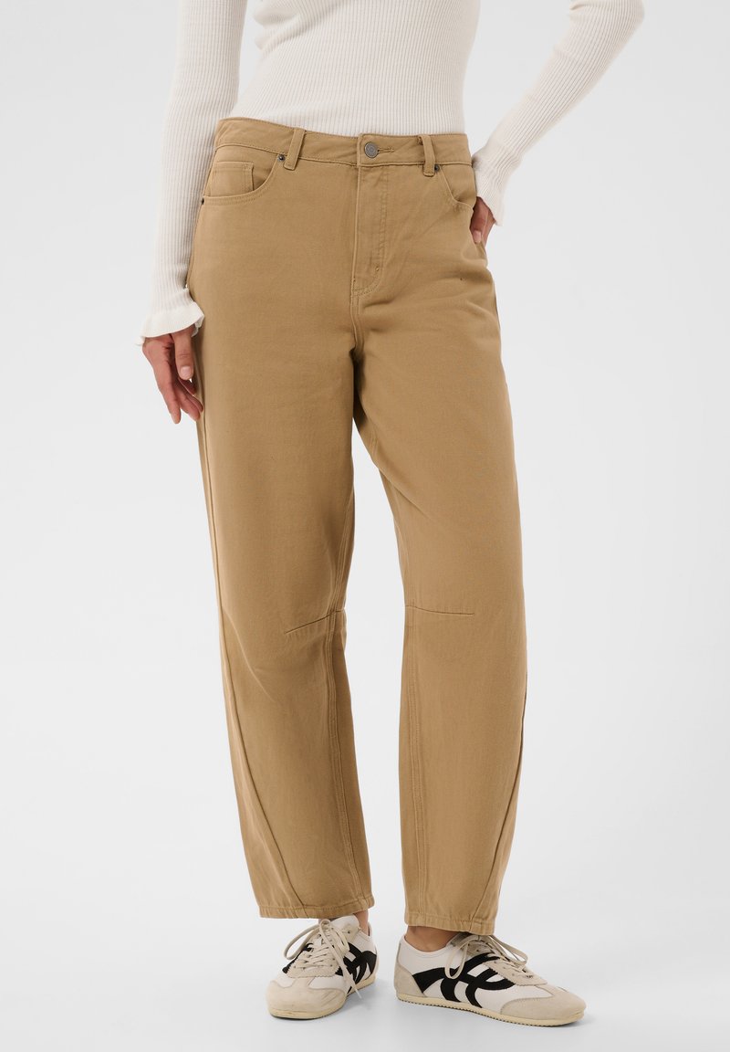 Personne portant un pantalon beige à jambes droites, un haut blanc à manches longues côtelé et des baskets blanches avec des accents noirs, debout devant un fond uni.