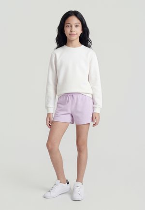 Κορίτσι που στέκεται φορώντας λευκό φούτερ με μακριά μανίκια, λιλά σορτς και λευκά αθλητικά παπούτσια μπροστά από μια απλή ανοιχτή επιφάνεια.
