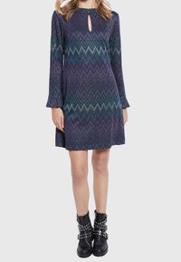 Abito a maniche lunghe con dettaglio a chiave, caratterizzato da un motivo a zigzag in tonalità di viola e verde petrolio. Indossato con stivali alla caviglia neri decorati con borchie.