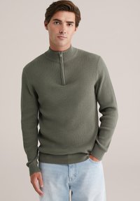 Jonge man in een olijfgroene geribbelde trui met een kwart-rits en lichtblauwe jeans, staand met één hand in de zak.
