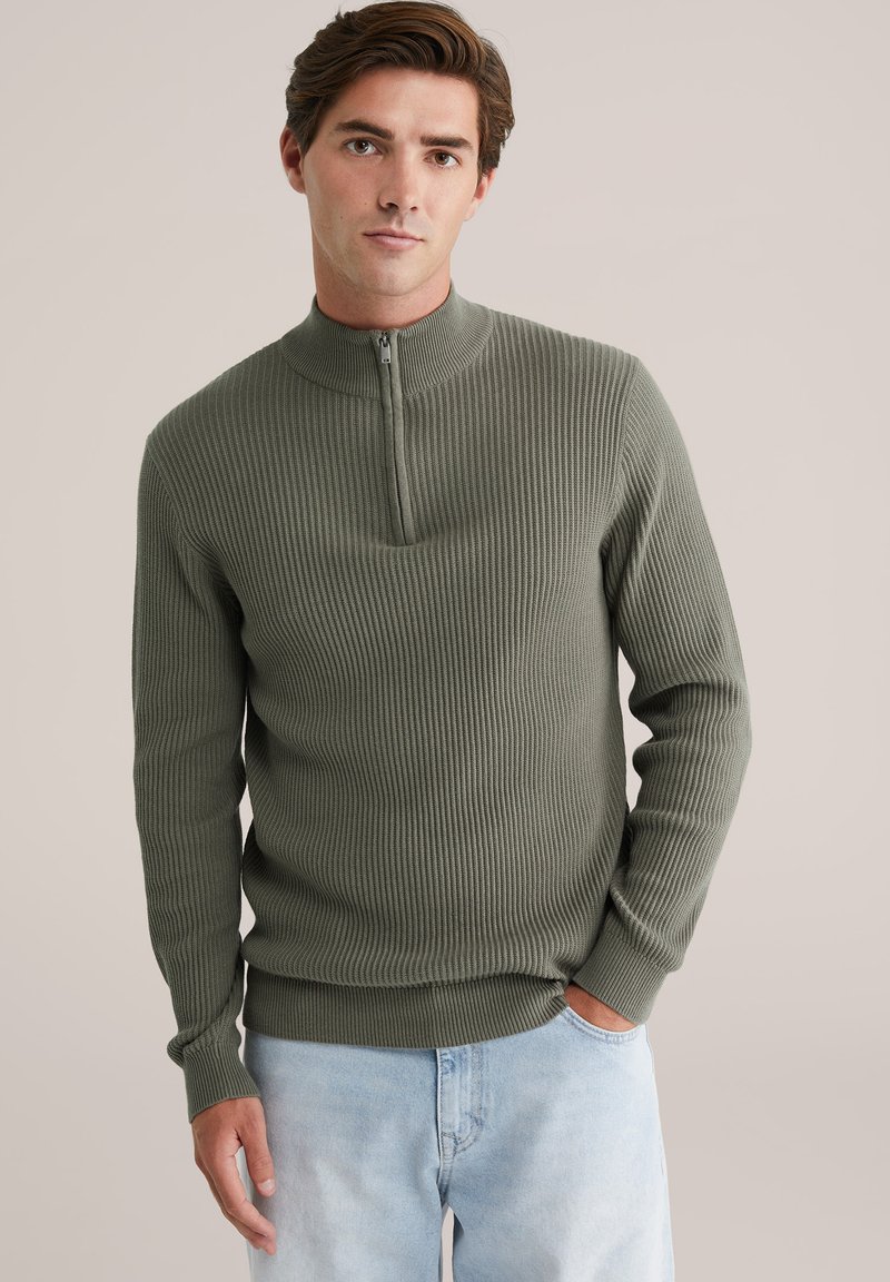 Jonge man in een olijfgroene geribbelde trui met een kwart-rits en lichtblauwe jeans, staand met één hand in de zak.