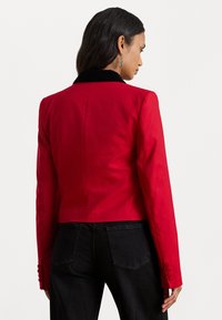 Červená skrátená bunda s čiernym zamatovým golierom, hladká tkanina, dlhé rukávy a gombíkové detaily. Nosená cez čierne džínsy, ktoré ukazujú priliehavý strih.