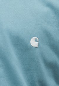 Lichtblauwe stof met een klein wit geborduurd logo in de vorm van een afgeronde, gestileerde "C" nabij het midden.