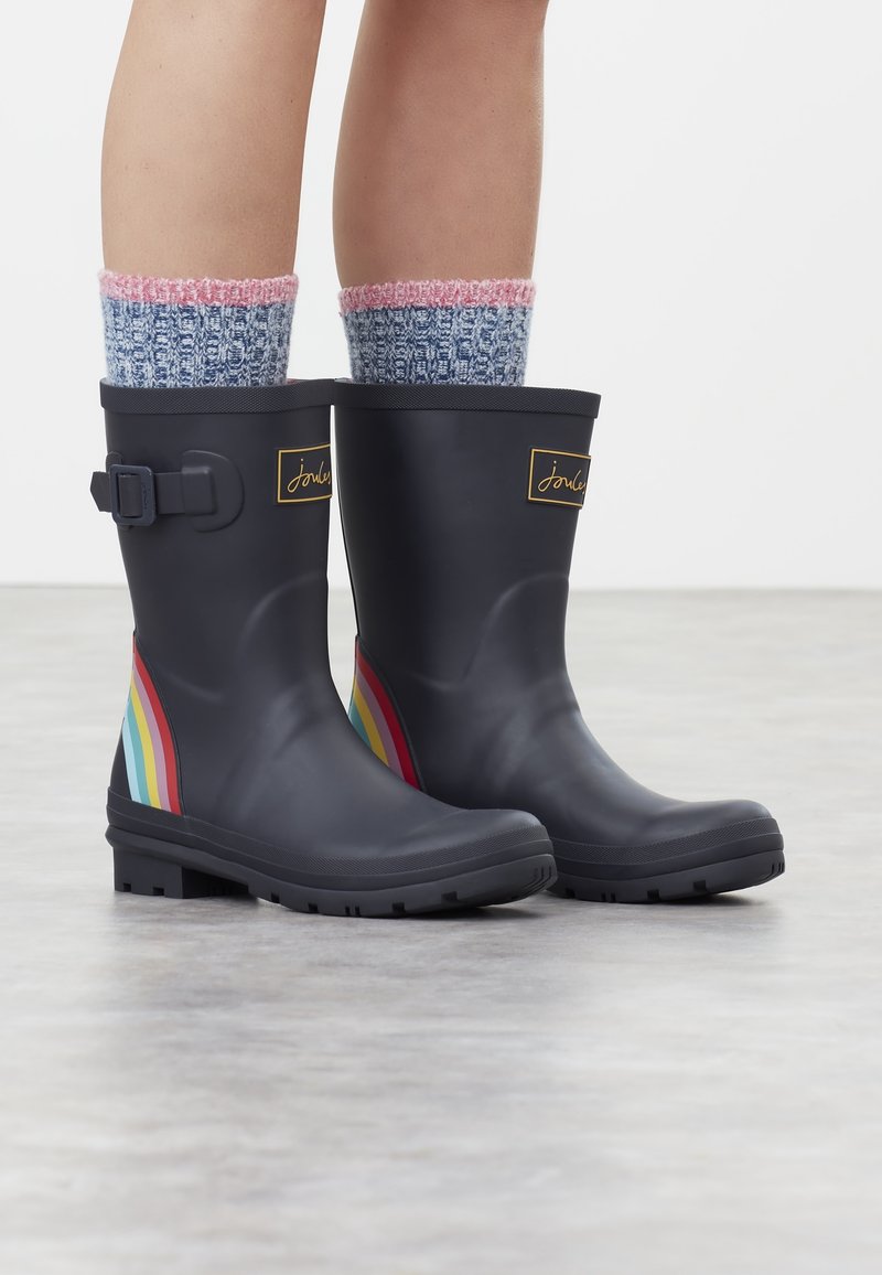 Svarta gummistövel med spänne, med flerfärgat randigt mönster och texturerad gummisula. Bärs med blåa stickade strumpor.
