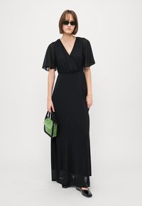 Black maxidress with short flutter sleeves, wrap design, and smooth fabric. Model holds a small green textured handbag and wears sunglasses.