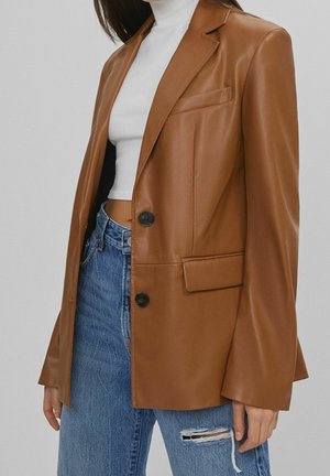 Donna che indossa un blazer in pelle marrone sopra un dolcevita bianco corto e jeans blu strappati, in piedi davanti a uno sfondo semplice.