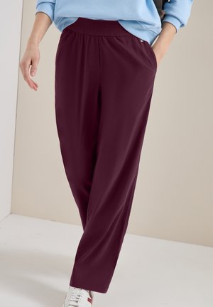 Personne portant un pantalon bordeaux foncé coupe décontractée et des baskets blanches, avec une main dans la poche, sur un fond uni.