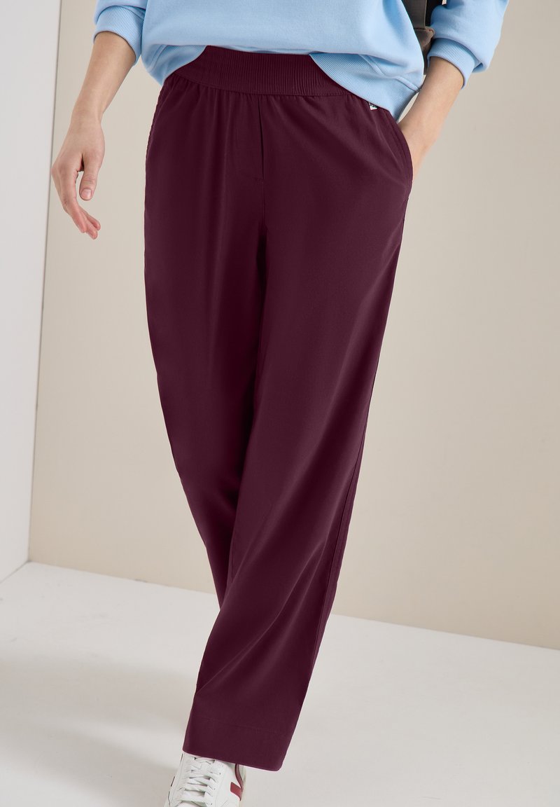Personne portant un pantalon bordeaux foncé coupe décontractée et des baskets blanches, avec une main dans la poche, sur un fond uni.