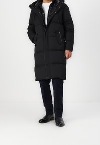 Man i en lång svart bubbelfodrad jacka med dragkedjefickor, marinblå byxor och svarta läderankelboots, stående mot en enkel vit bakgrund.