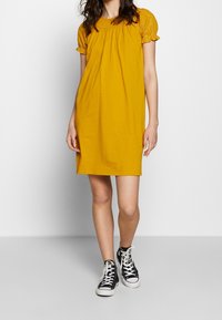 Abito giallo senape con maniche a sbuffo in pizzo corte, scollatura arricciata e vestibilità comoda, abbinato a sneakers nere alte.