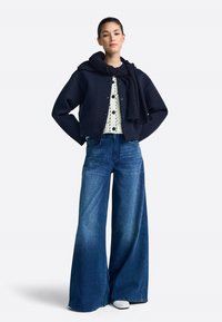 Marineblauw cropped jasje over een wit shirt met stippen, gecombineerd met wijd uitlopende blauwe jeans en grijze schoenen; gestructureerde stof en een relaxte pasvorm.