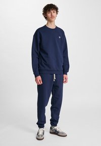 Ung man med lockigt hår som bär en marinblå sweatshirt och matchande sweatpants, stående mot en ljus enfärgad bakgrund.