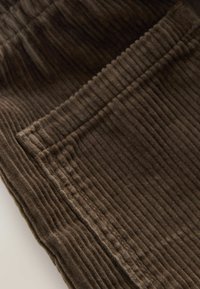 Braunes Cordstoffgewebe mit vertikalen Streifen, versehen mit einer Naht und einem Taschen Detail, das eine weiche Textur und eine satte Farbe hervorhebt.