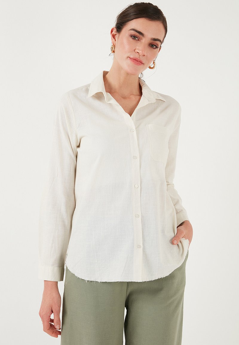 Camicetta bianca in lino con chiusura a bottoni, maniche lunghe, taschino sul lato sinistro del petto e orlo curvo, indossata con pantaloni verde chiaro.