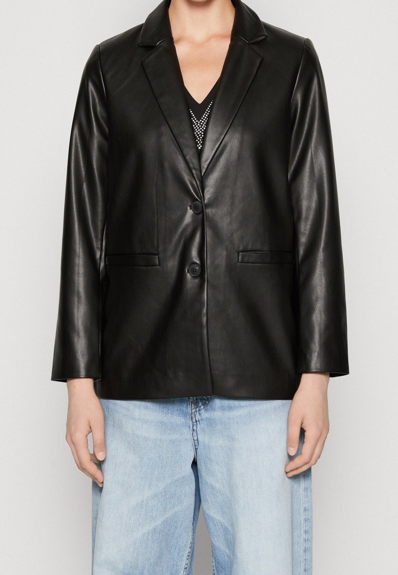 Personne portant un blazer en cuir noir à deux boutons sur un jean bleu clair et un haut noir avec des clous argentés.