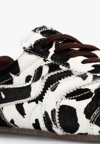 Zapatilla de cuero sintético con estampado de vaca en blanco y negro, parte superior con textura de pelaje, cordones marrones, detalles de costuras plateadas y suela de goma marrón.