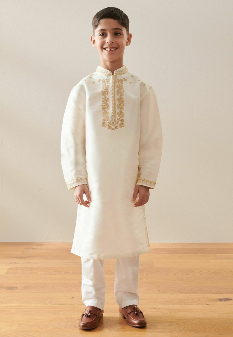 Garçon debout à l'intérieur portant une tenue traditionnelle crème avec broderies dorées sur le col et la poitrine, pantalon blanc et chaussures marron, souriant.